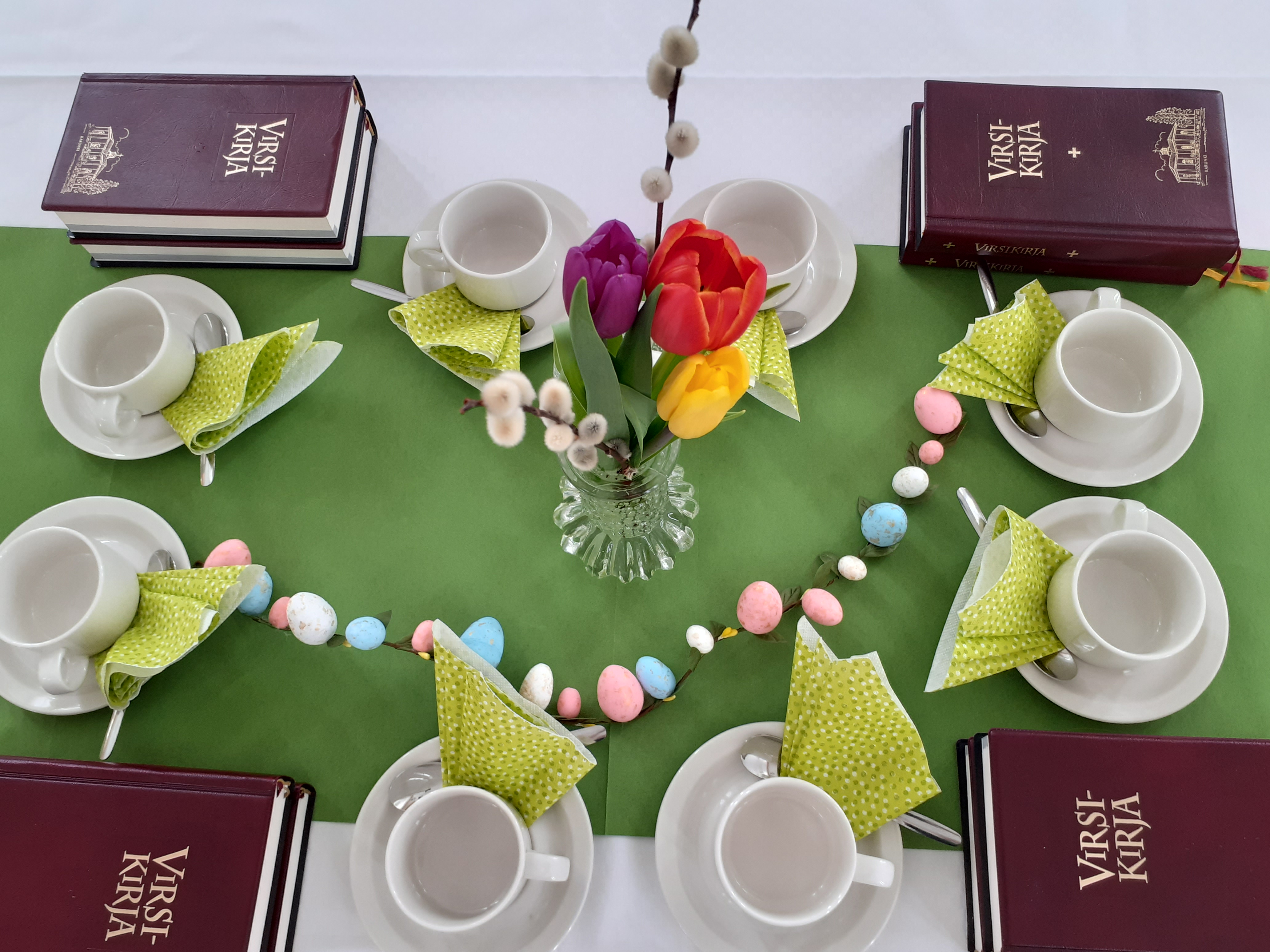 Katettu pöytä kahvikupeilla, tulppaaneilla, virsikirjoilla ja pääsiäiskoristeilla.