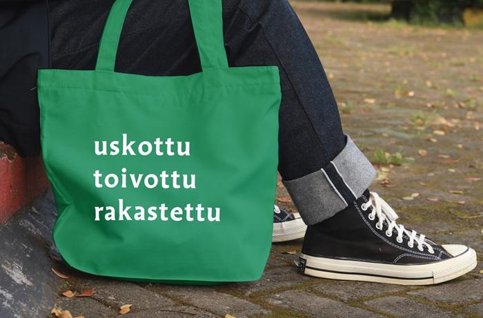 Vihreä kassi, jossa tekstinä: uskottu, toivottu, rakastettu. Kassin kantajalla tummat farkut ja tennarit.